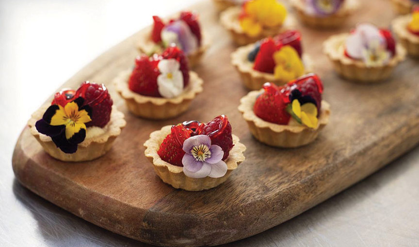 Miniature shortcrust pastry case canapes topped with glazed strawberries and pansies sitting atop a rustic wooden board