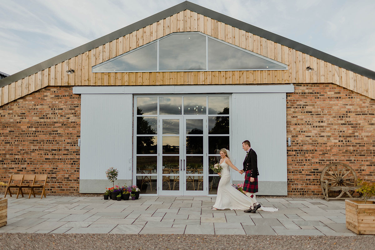 A bride and groom holding hands walking past a stone barn with blue doors