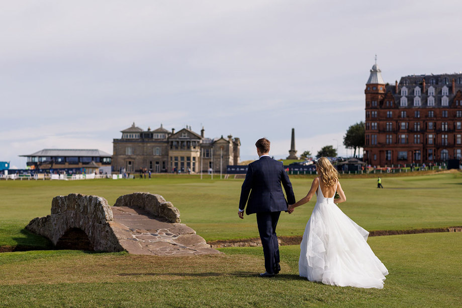 A couple in wedding attire holding hands and walking across the Old Course with elegant buildings in the background, captured by Duke Photography