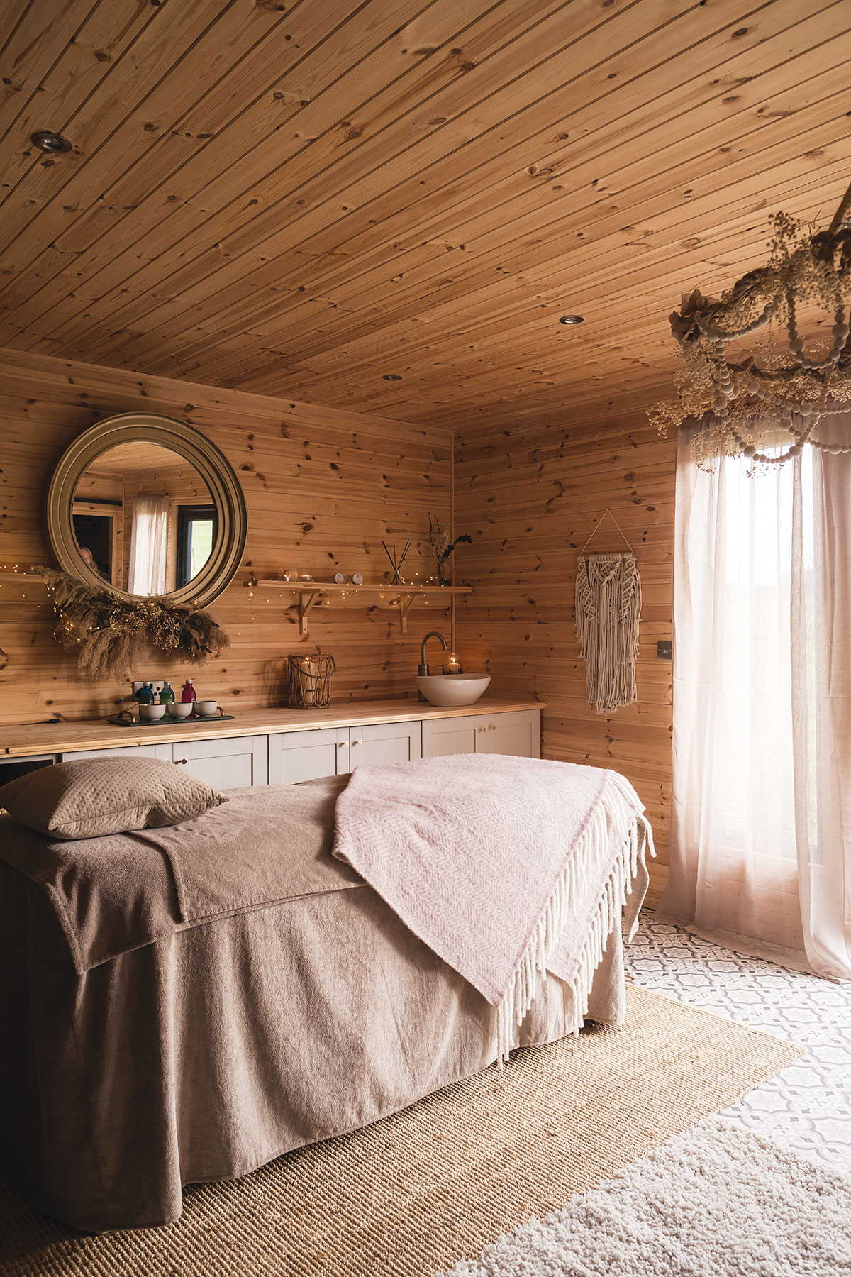 a wooden walled and ceiling room has a tall single bed in the middle, covered in plenty of towels and a blanket and behind the bed is a counter with a sink and bowls of products with a mirror hanging above