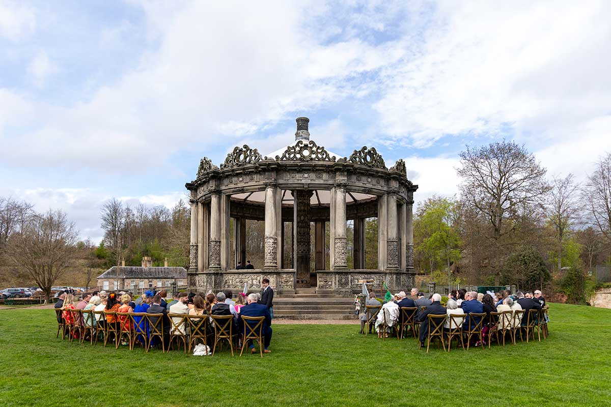 Outdoor wedding ceremony at Restoration Yard in Dalkeith Country Park with guests seated around a historic stone pavilion.