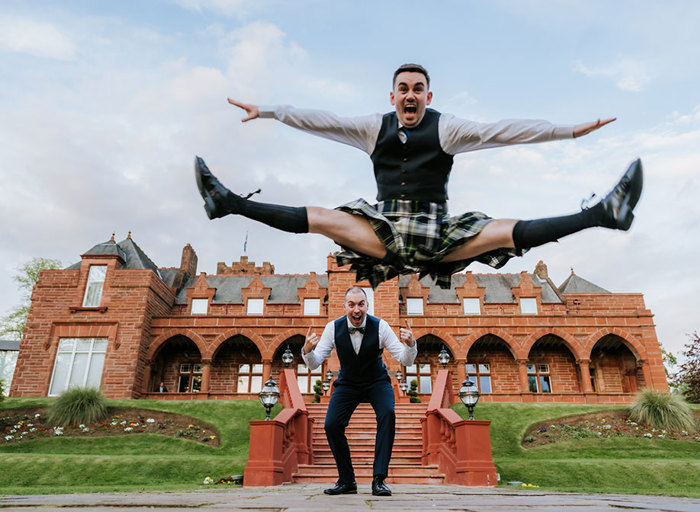 a groom in a kilt jumps above another groom wearing a suit outside a red sandstone building 