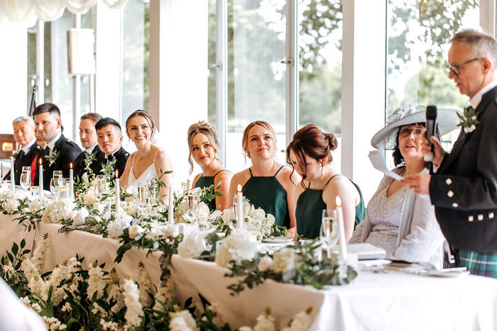 bride, groom and bridesmaids look towards father of the groom while sitting at long table with foliage at mar hall