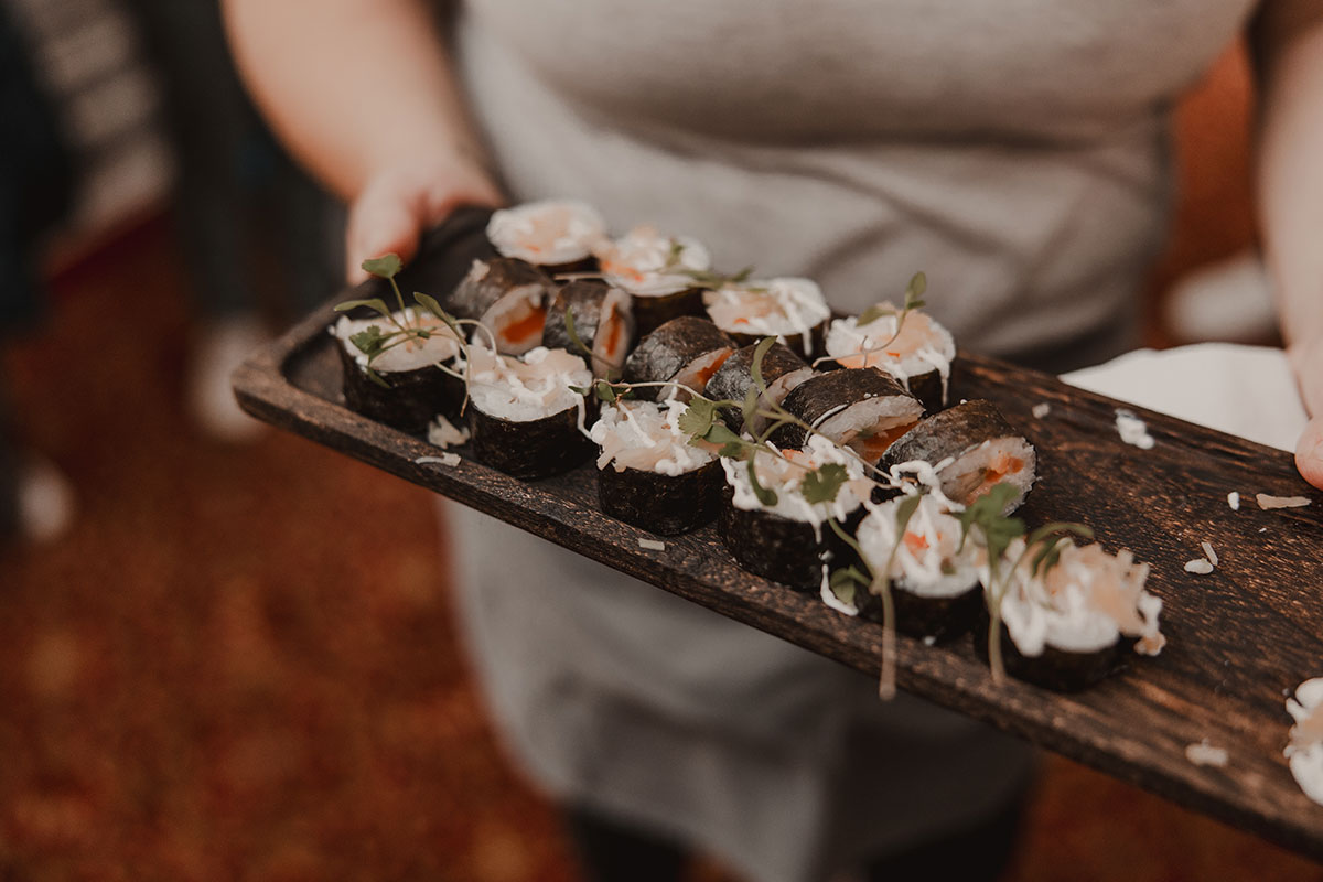 A server holding a rustic wooden platter of sushi rolls garnished with micro herbs, ready to be served at a wedding reception