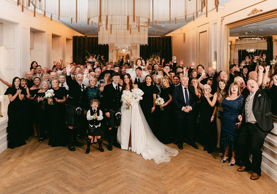 full room of a wedding group photo where brunette bride in high leg slit wedding dress with long puff sleeves and groom in full dark tartan kilt outfit stand in the centre with all others around them, holding their hands up in the air