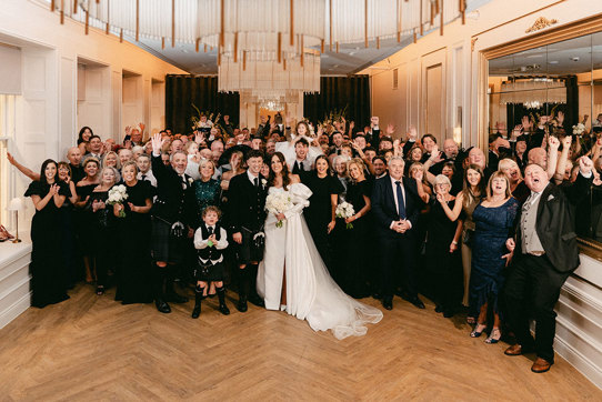 full room of a wedding group photo where brunette bride in high leg slit wedding dress with long puff sleeves and groom in full dark tartan kilt outfit stand in the centre with all others around them, holding their hands up in the air