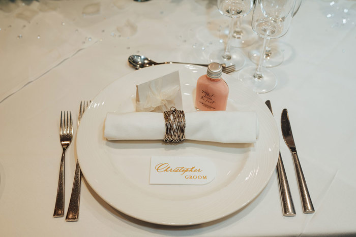 a detail of a table setting at a wedding featuring a white plate with silver napkin holder, a small bottle, and groom place name setting
