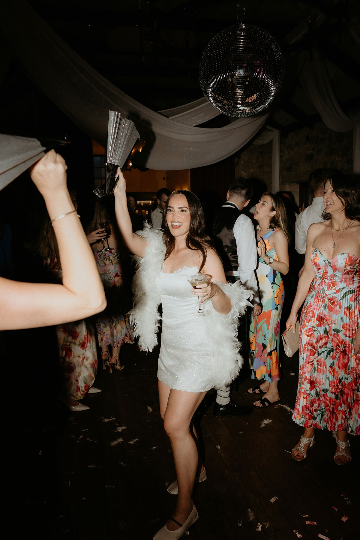 Bride dancing in feather-trimmed mini reception dress under disco ball at Rosebery Steading wedding party in Midlothian.