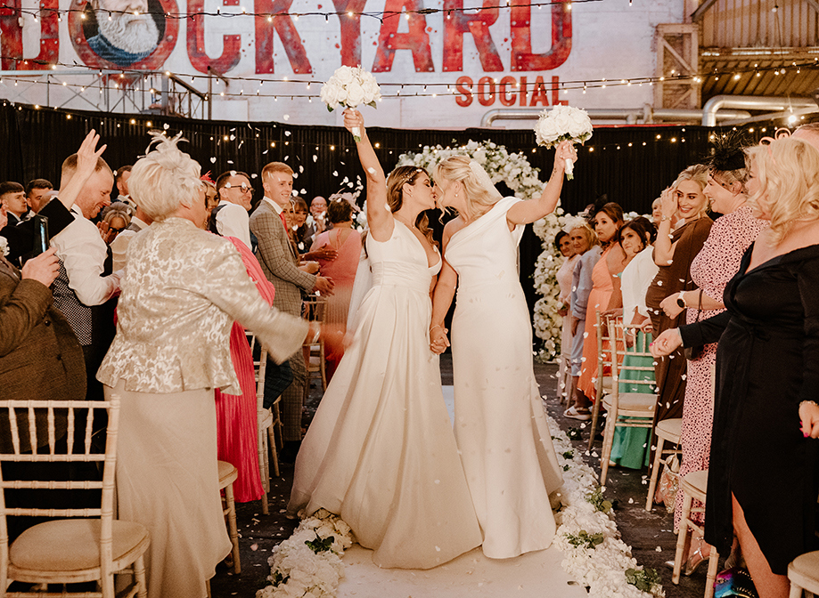 two brides kiss and hold their bouquets aloft while standing on the aisle after their Dockyard Social wedding ceremony. Guests cheer and throw confetti either side