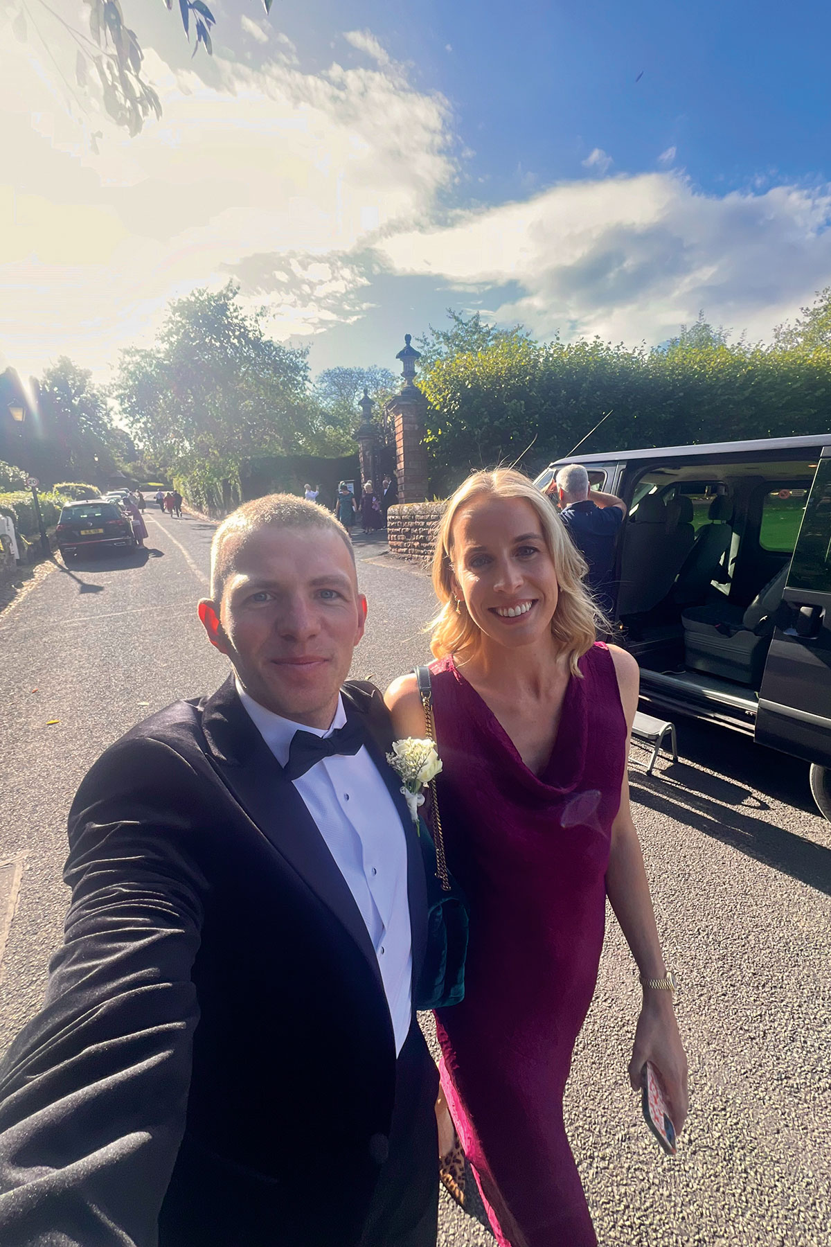 Couple taking a selfie on a sunny street beside a parked car; the man wears a black tuxedo with boutonniere and the woman wears a sleeveless burgundy dress