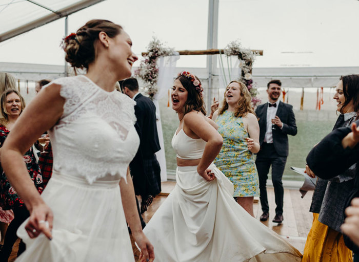 two brides dancing with wedding guests looking on. They are in a marquee with clear panel that offers a view to the grass outside