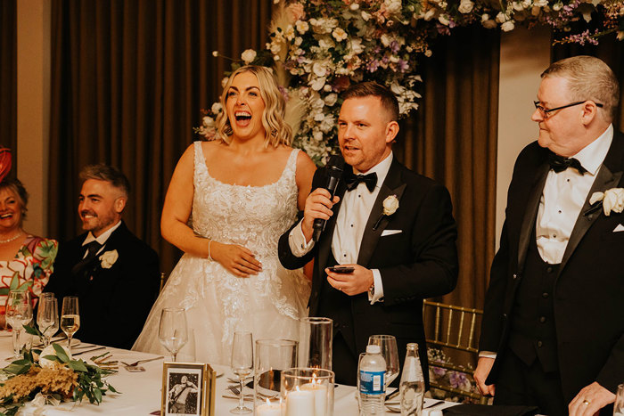 A bride standing next to a groom who is speaking into a microphone.