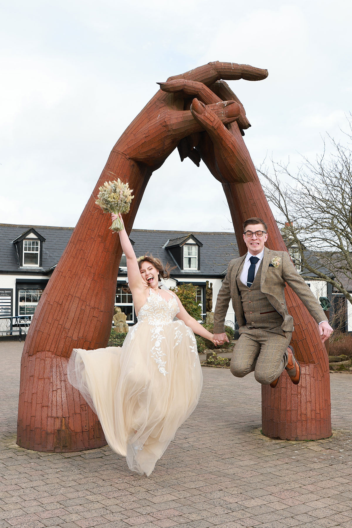 groom and bride holding bouquet jump in the air underneath hand sign at gretna green