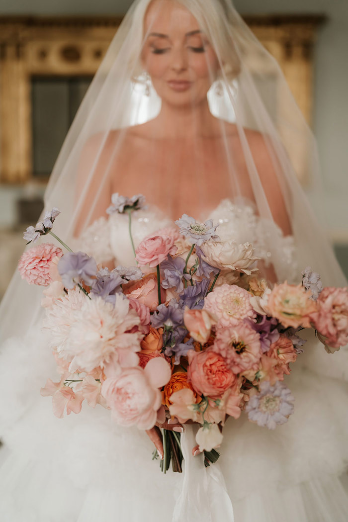 A bride holding a pink and purple bouquet with her veil over her face 