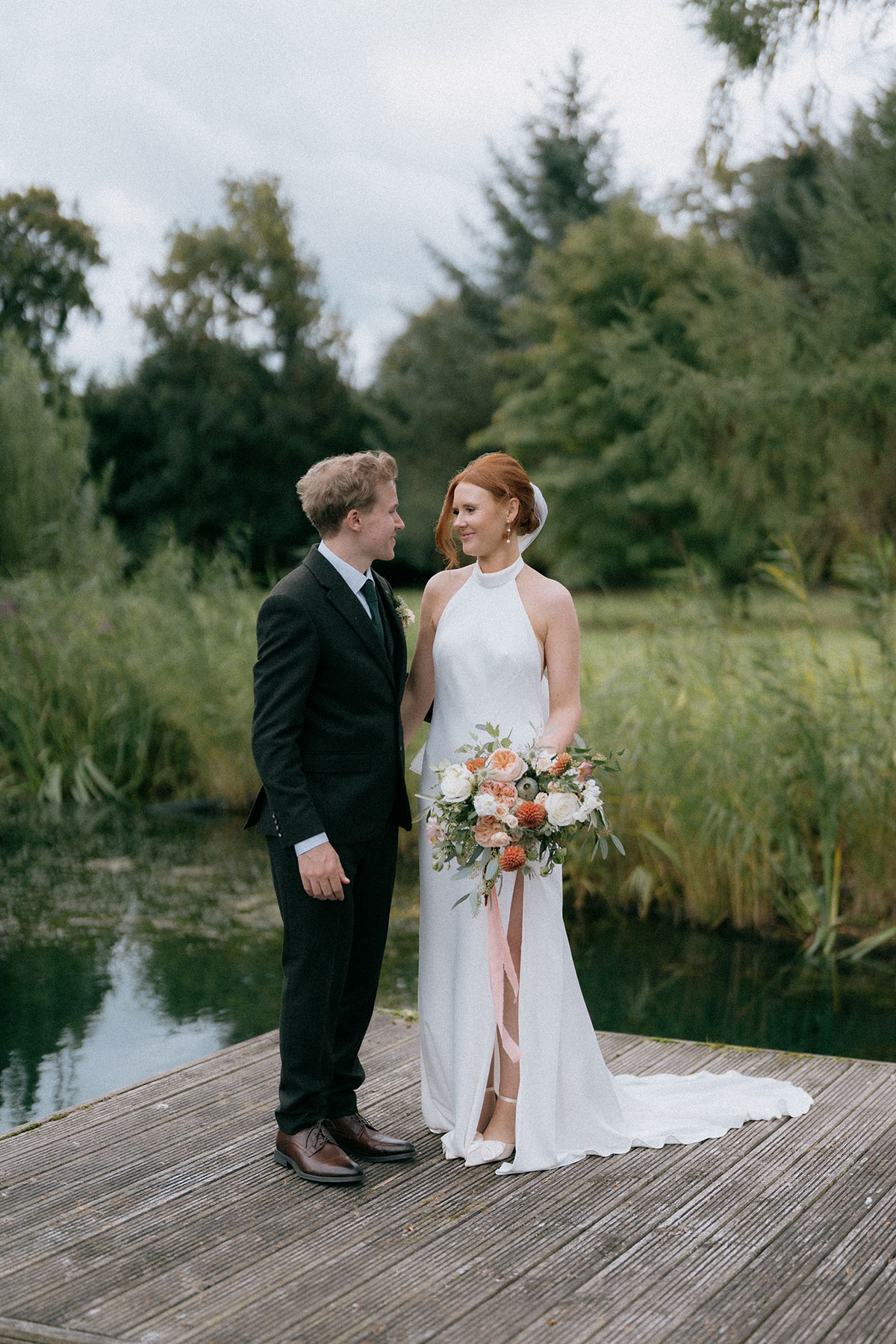Bride and groom standing together on lakeside jetty holding bouquet at countryside wedding