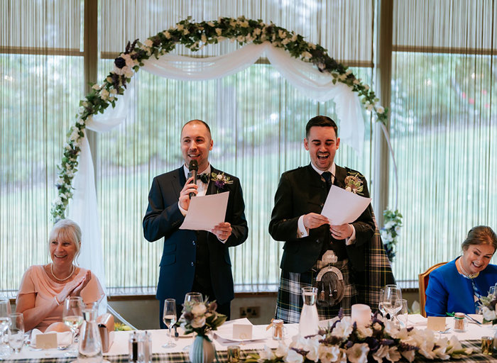 two grooms standing behind a long table holding sheets of paper and microphones with floral arch in background