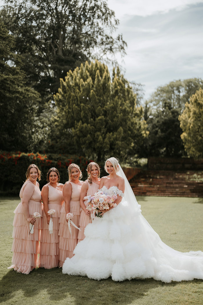 A bride wearing a wedding dress with a large skirt standing next to four bridesmaids wearing pink dresses 