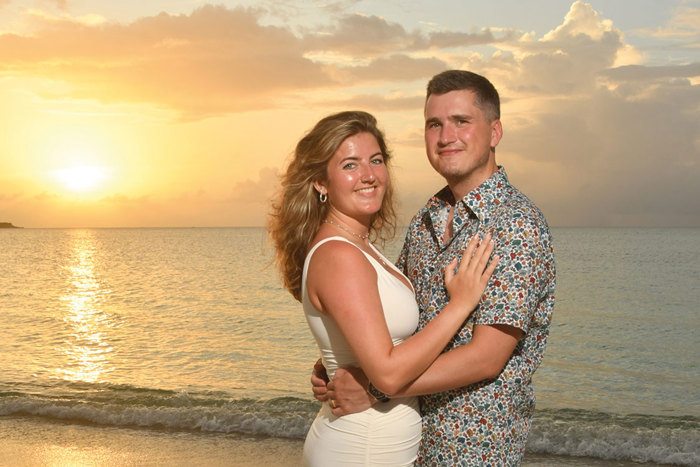 couple with arms wrapped around each other, stood in front of beach sunset