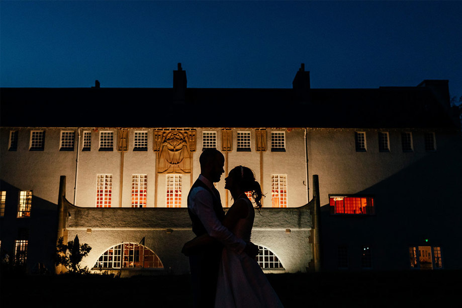 A couple looking at each other are backlit against a white building which is lit up at night