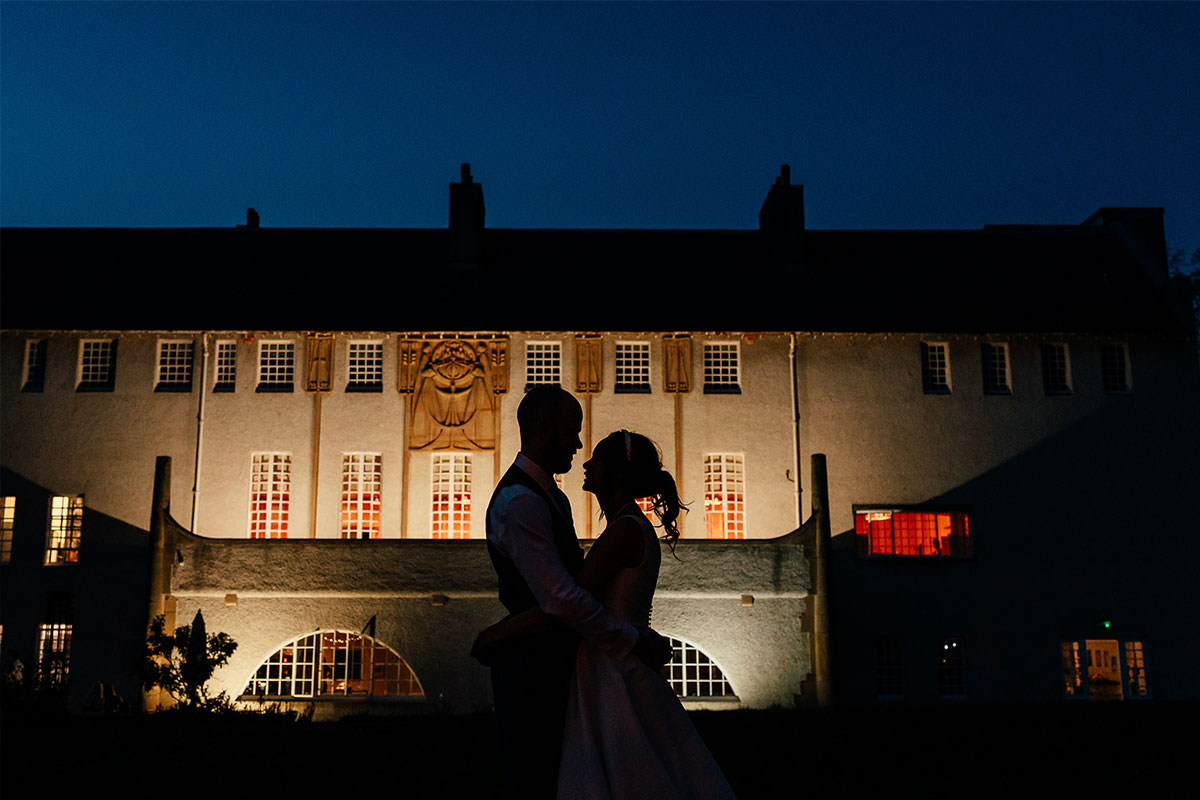 A couple looking at each other are backlit against a white building which is lit up at night