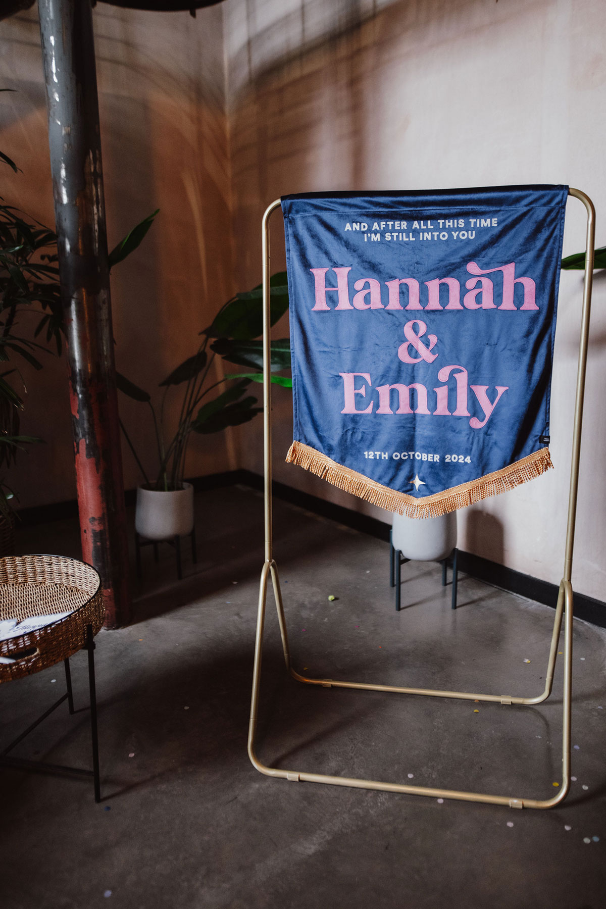 Fabric wedding banner displayed on a gold stand reading ‘Hannah & Emily’ with a Paramore lyric, styled as part of the Engine Works décor.