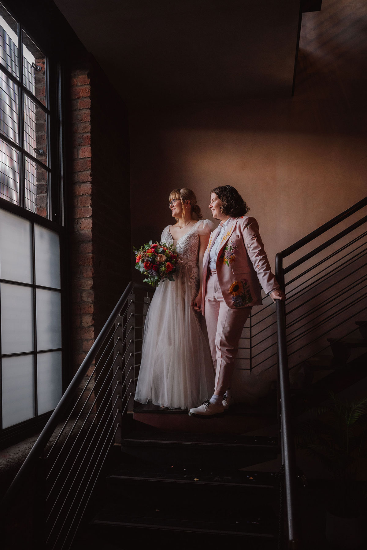 Two brides posing on the Engine Works staircase, looking out the window with soft industrial lighting behind them.