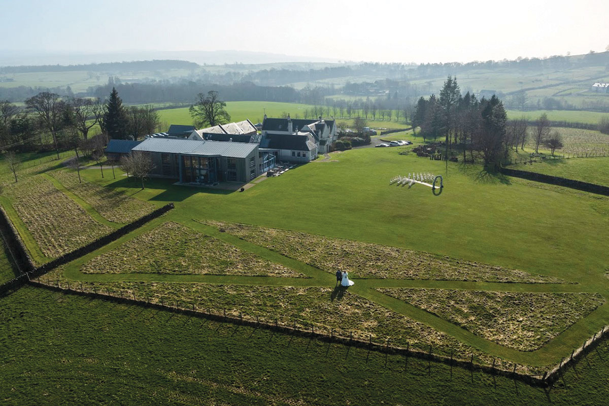 overhead view of bride and groom stood out on grassy field, in the centre of a Saltire marked into the ground