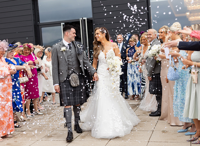 a bride and groom walking through two rows of guests throwing confetti. They are outside a black wooden-clad building with large glass windows