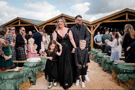 Joyous Bride And Groom Wearing Black Walking Up An Aisle Surrounded By Rows Of Hay Bales And Holding Hands With Two Children Wearing Black