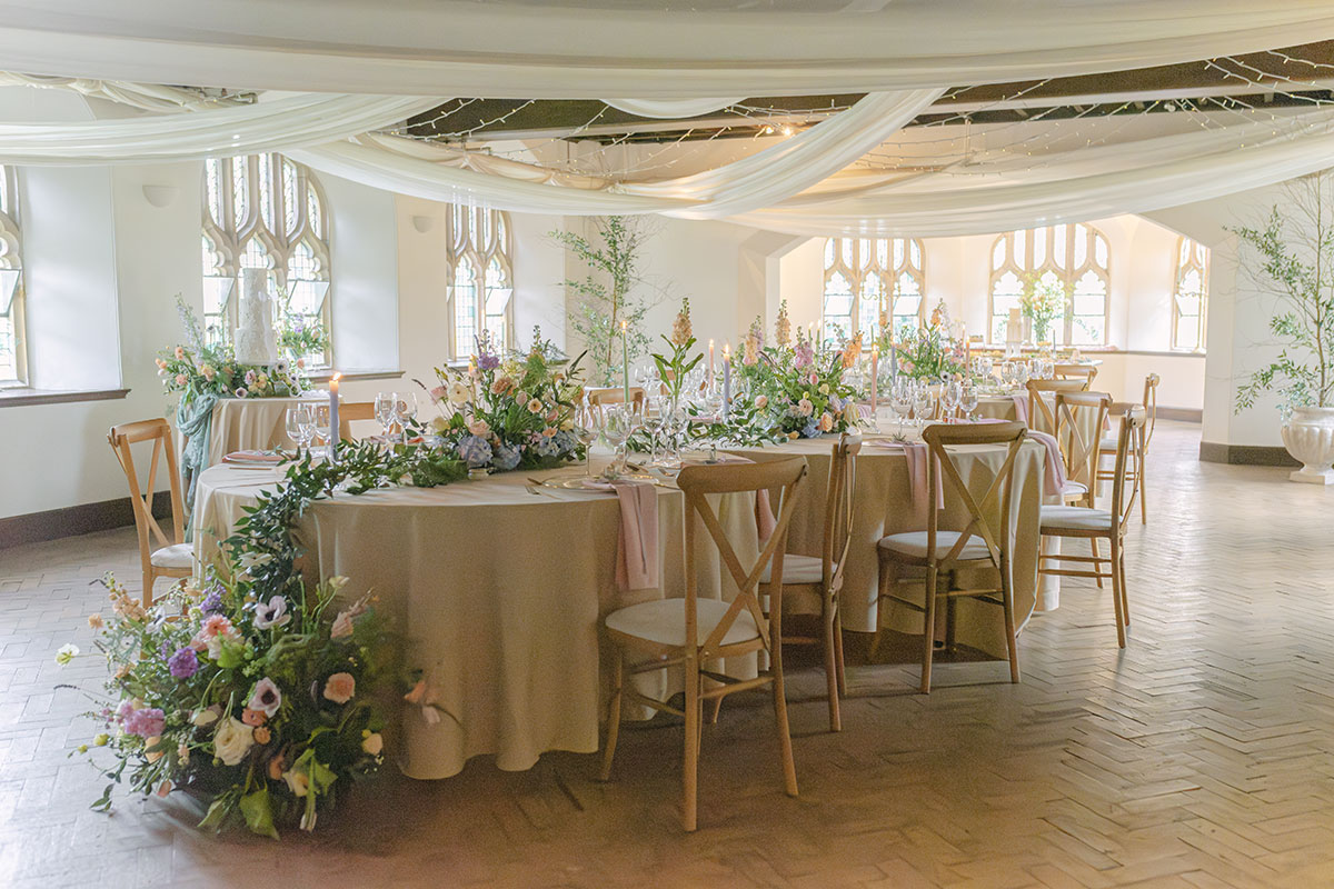 Elegant wedding reception setup with round tables, floral centrepieces, draped ceiling fabric, and candles in a bright room