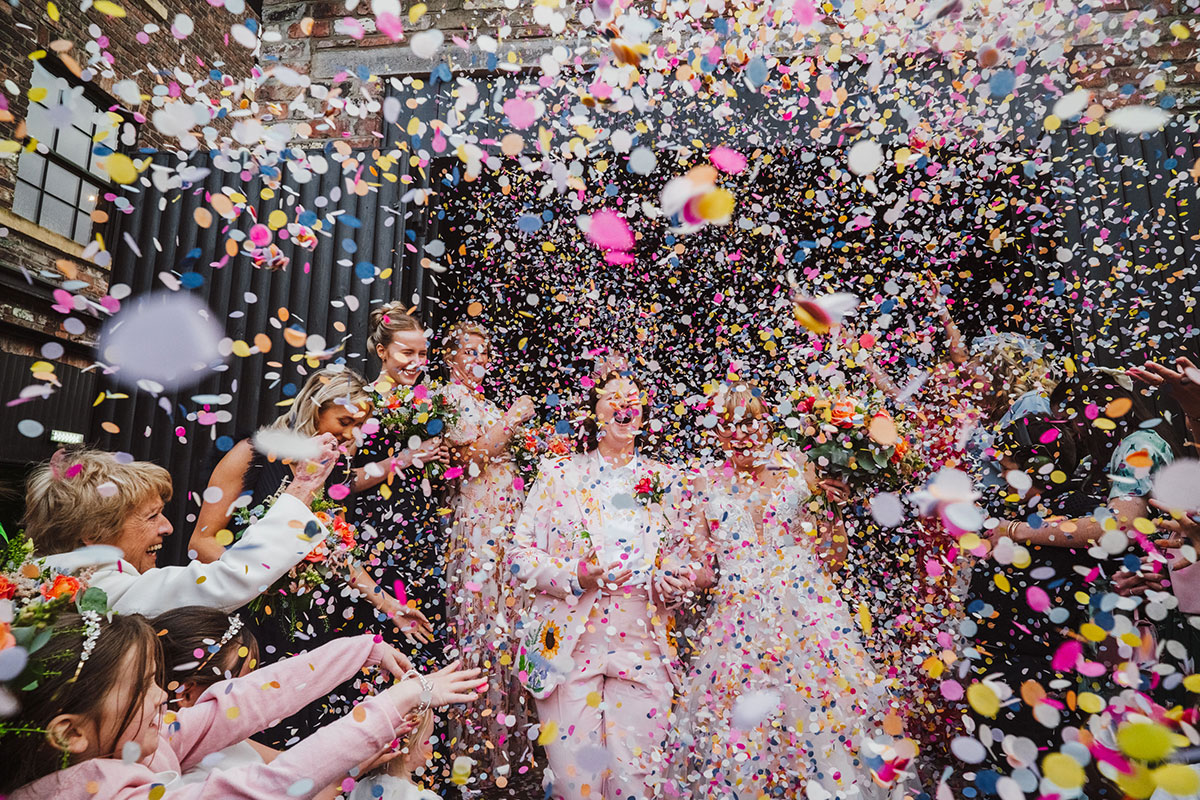 Close-up moment of the brides laughing as a huge burst of multicoloured confetti surrounds them during their Engine Works wedding celebration.