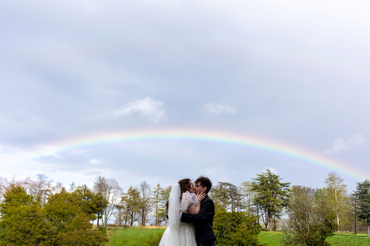 a bride and groom kissing underneath a rainbow with trees behind them
