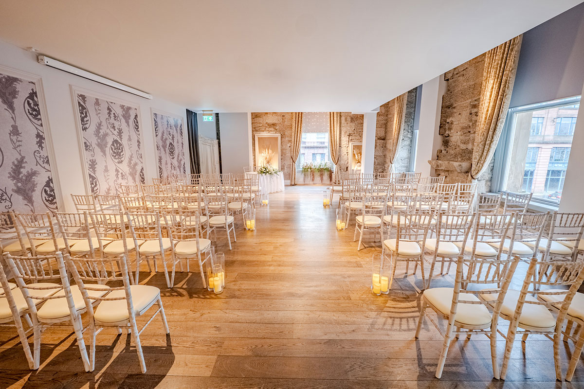 A bright ceremony room with rows of Chiavari chairs, wooden flooring and soft candlelight along the aisle.