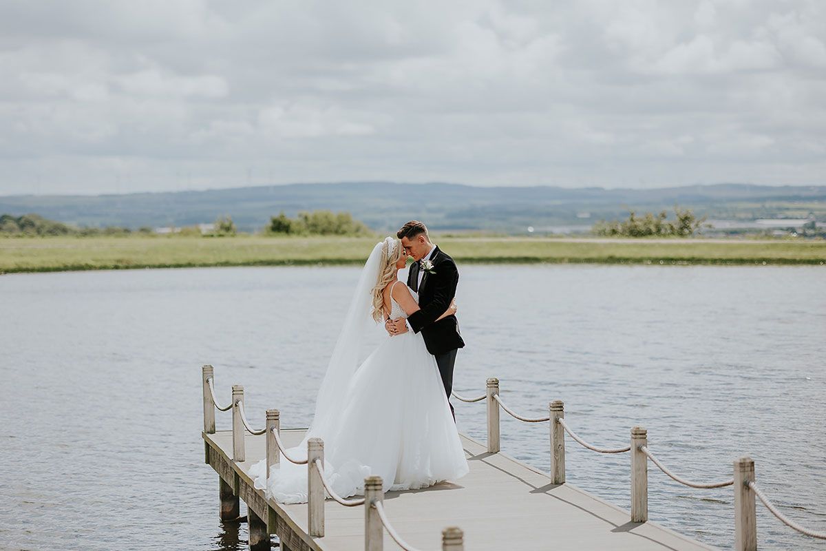 blonde bride in ballgown and groom in tux embrace, forehead to forehead on a dock with calm waters around them