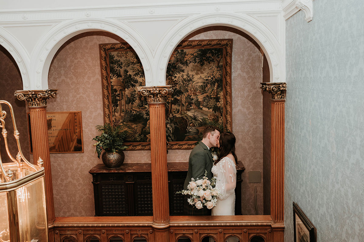 Couple kissing on a balcony inside Norton House, framed by grand architecture and warm lighting.