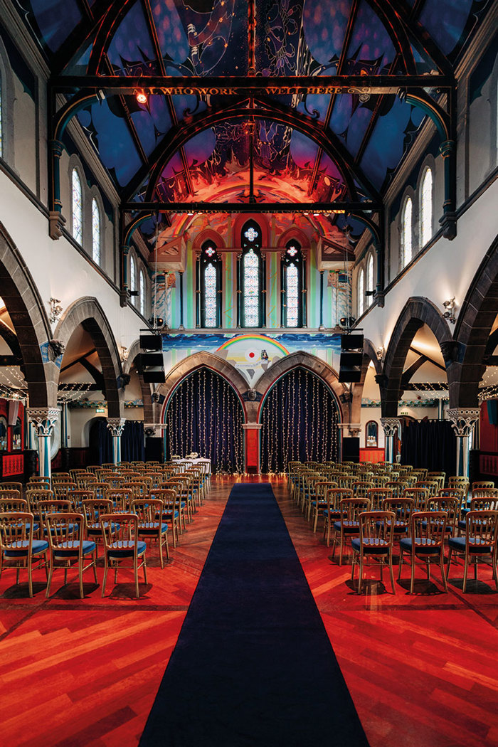 rows of identical chairs are lined up with a dark blue runner creating an aisle down the middle of them, the venue complete with tall ceilings, colourful sky mural and wide arches throughout the space
