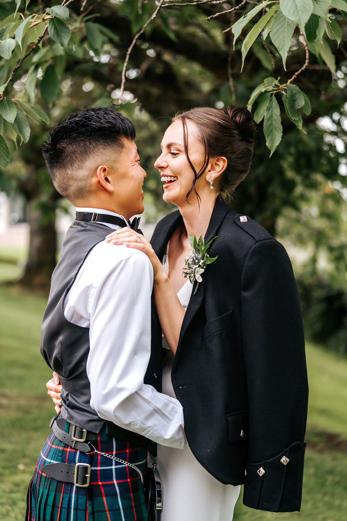 bride and groom smile at one another while bride wears gooms kilt jacket under tree