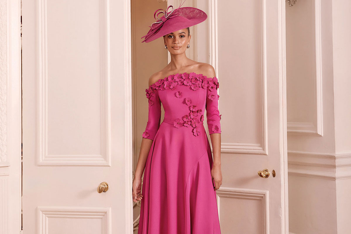 Pink off-the-shoulder mother of the bride dress with floral appliqué and matching statement hat.