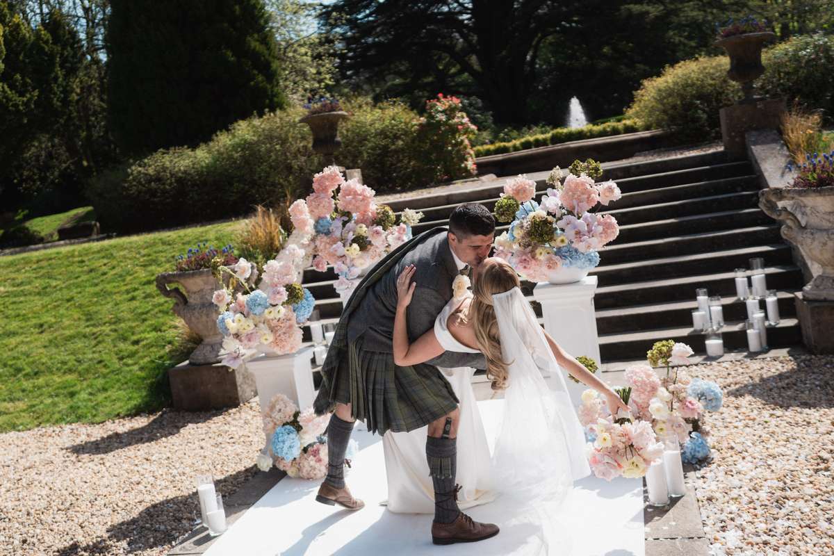 n the garden Alt text: Stuart dips Siân for a kiss surrounded by pastel pink and blue flowers at their outdoor Mar Hall wedding ceremony.