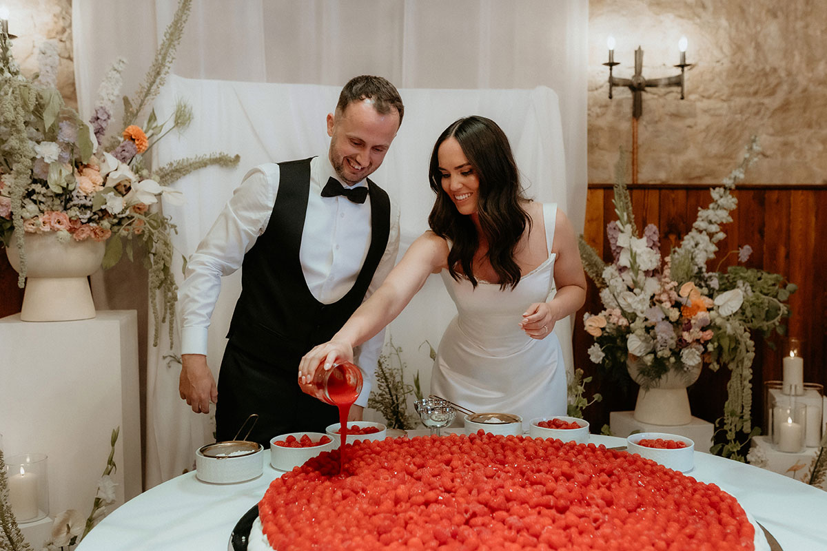Bride and groom pouring berry sauce over giant pavlova wedding dessert at Rosebery Steading reception in Midlothian.