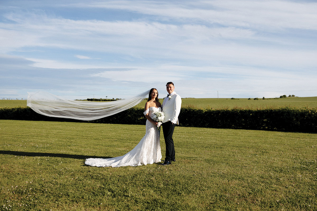 Bride and groom stand together on the grass at Falside Mill, the bride’s veil flowing out behind her in the countryside breeze.