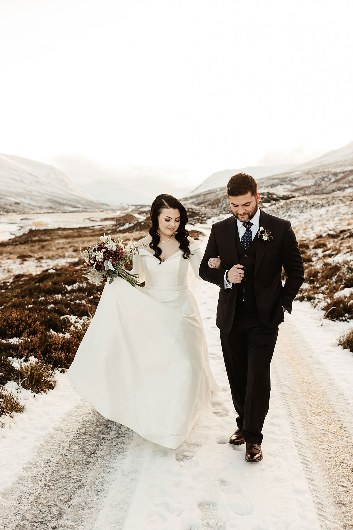 Bride holding her gown and bouquet as she walks arm in arm with the groom on a snowy track in the mountains