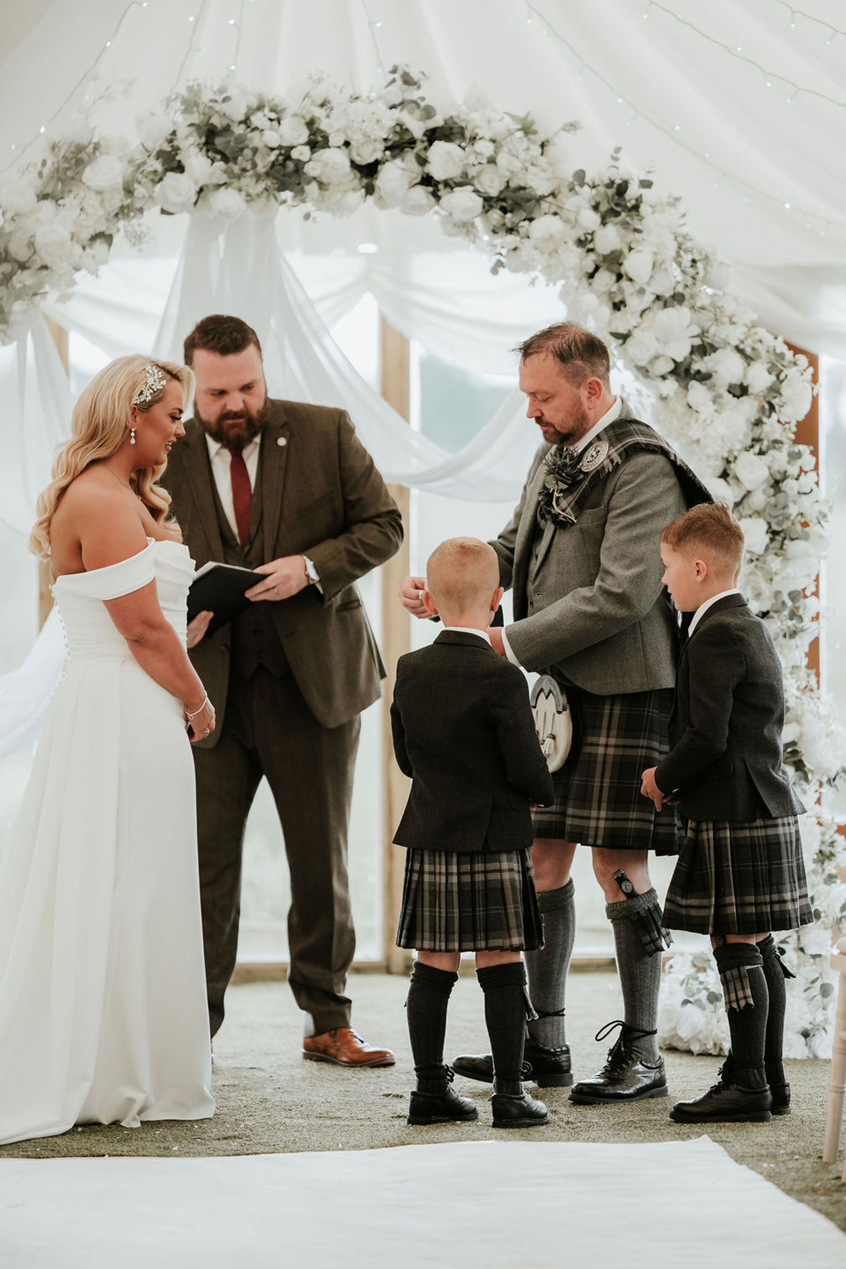 Couple stand with two boys and their celebrant beneath a white floral arch during the ceremony.