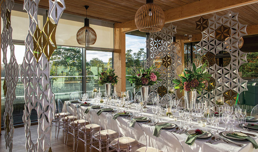 A long table surrounded by perspex chairs set for dinner with green napkins and wine glasses with view out through floor-to-ceiling windowsto lush green countryside