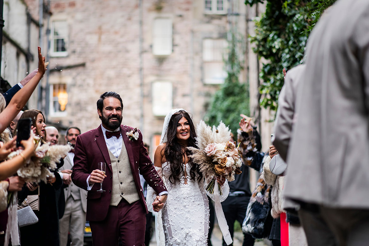 bride and groom, covered in remnants of confetti, hold hands as they walk outdoors amongst friends and family