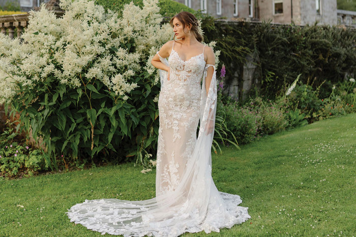 a brunette bride in a floaty floral embroidered wedding dress stands on a very green lawn of grass with a few white flower bushes in the background