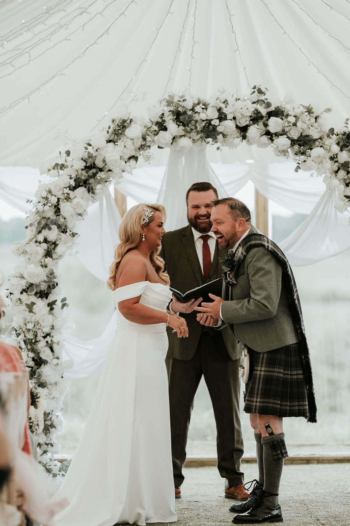 Bride and groom laughing together during the ceremony under a white floral arch.