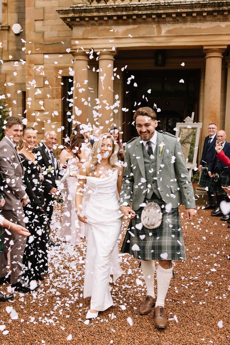 Newlyweds Lia and Louis smile as guests shower them with white confetti outside Cambo House in Fife.