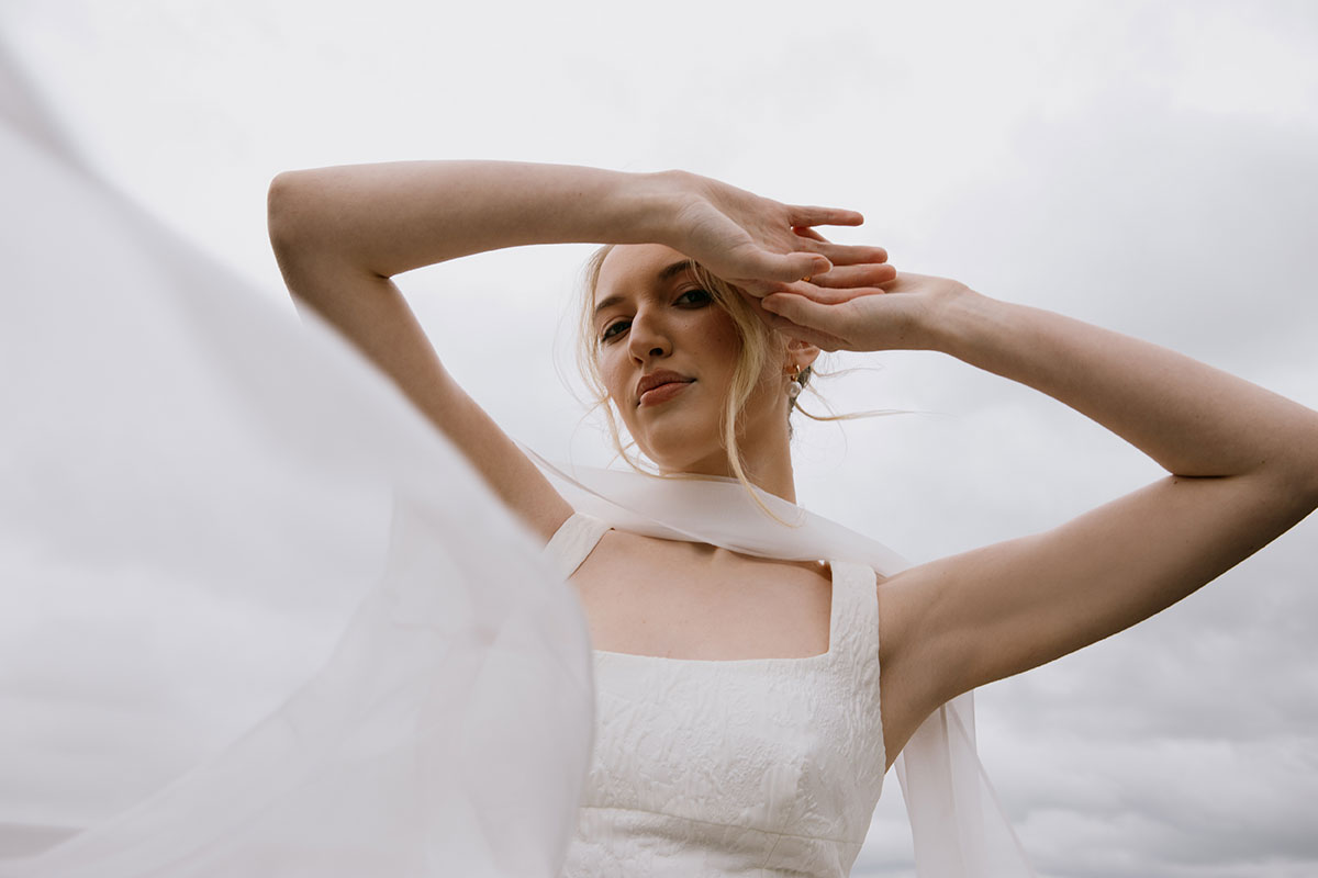 A bride in a square-neck textured gown poses with her arms lifted against a cloudy sky, with sheer scarf fabric flowing around her.