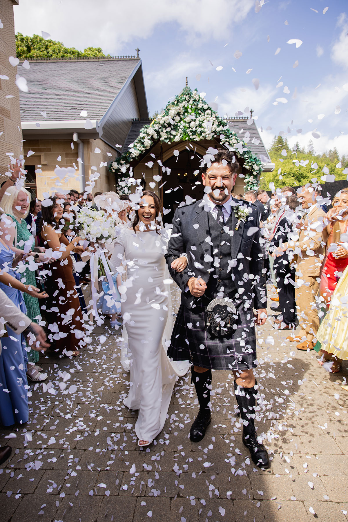 Bride and groom walk through a flurry of white petals outside the chapel surrounded by guests